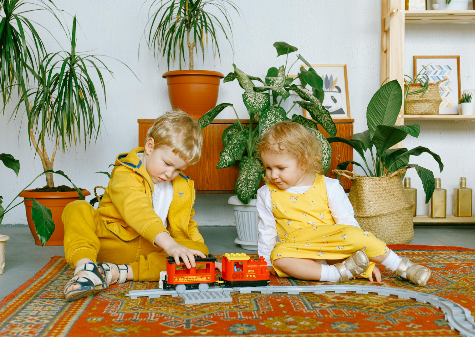 Two young children playing with a red toy train on a patterned rug in a living room with potted plants