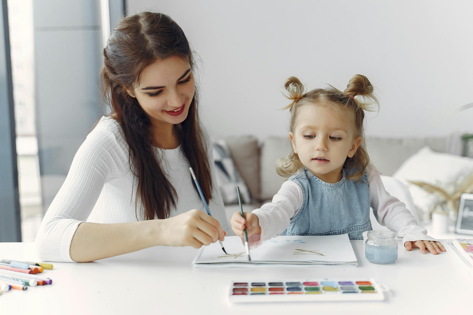 Woman helping a toddler paint with watercolors at a bright table in a living room