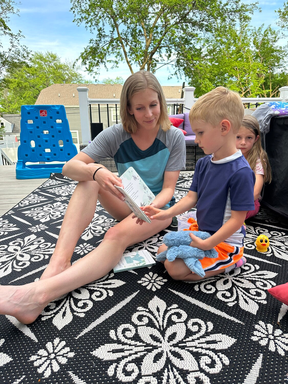 Woman reading a board book about emotions to a young boy on a patterned outdoor rug while another child watches