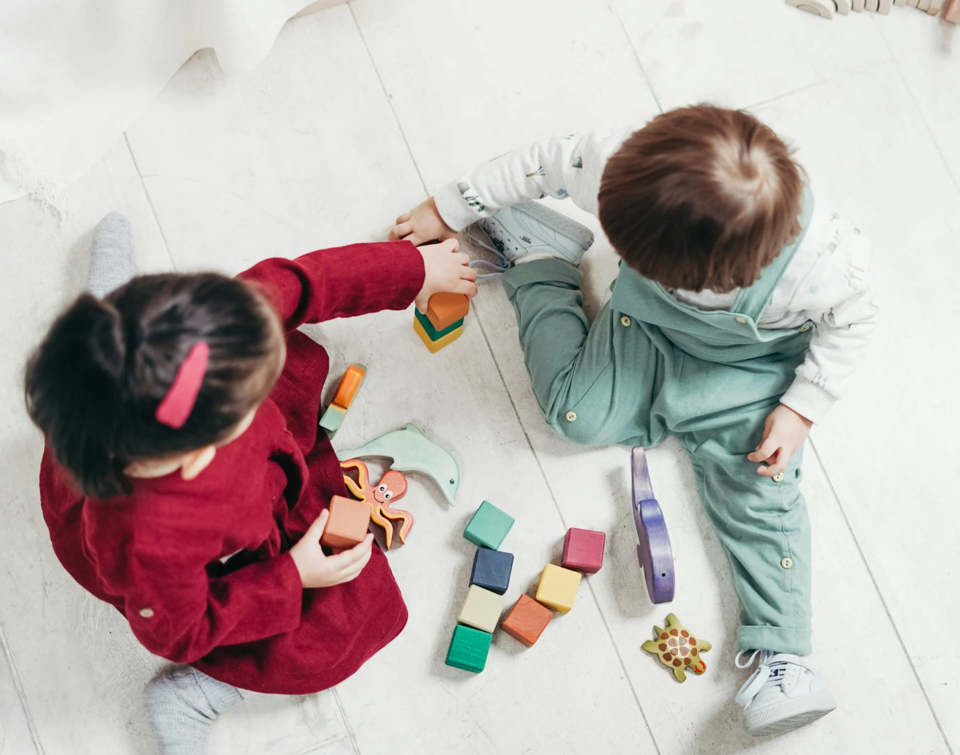 Two young children sitting on a light floor playing with colorful wooden blocks and animal toys