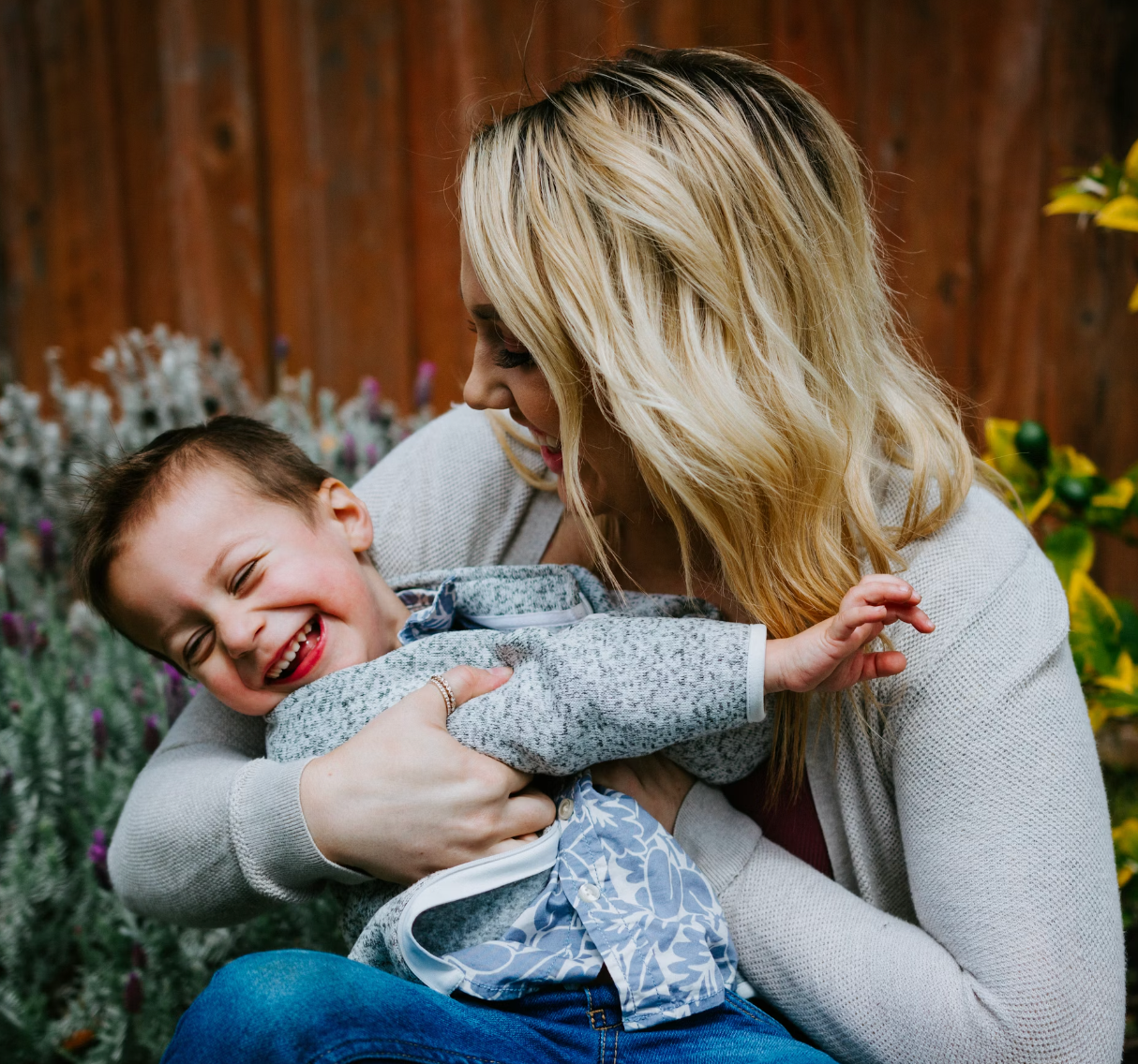 Mother hugging and laughing with her toddler outdoors in front of a wooden fence and lavender plants
