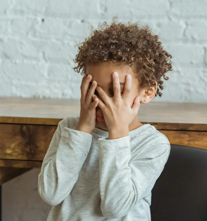 Child covering their face with both hands while sitting at a wooden table against a white brick wall, appearing anxious