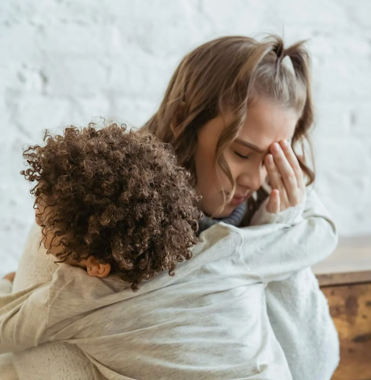 Parent holding child in a tight hug while covering their face in stress against a light brick wall
