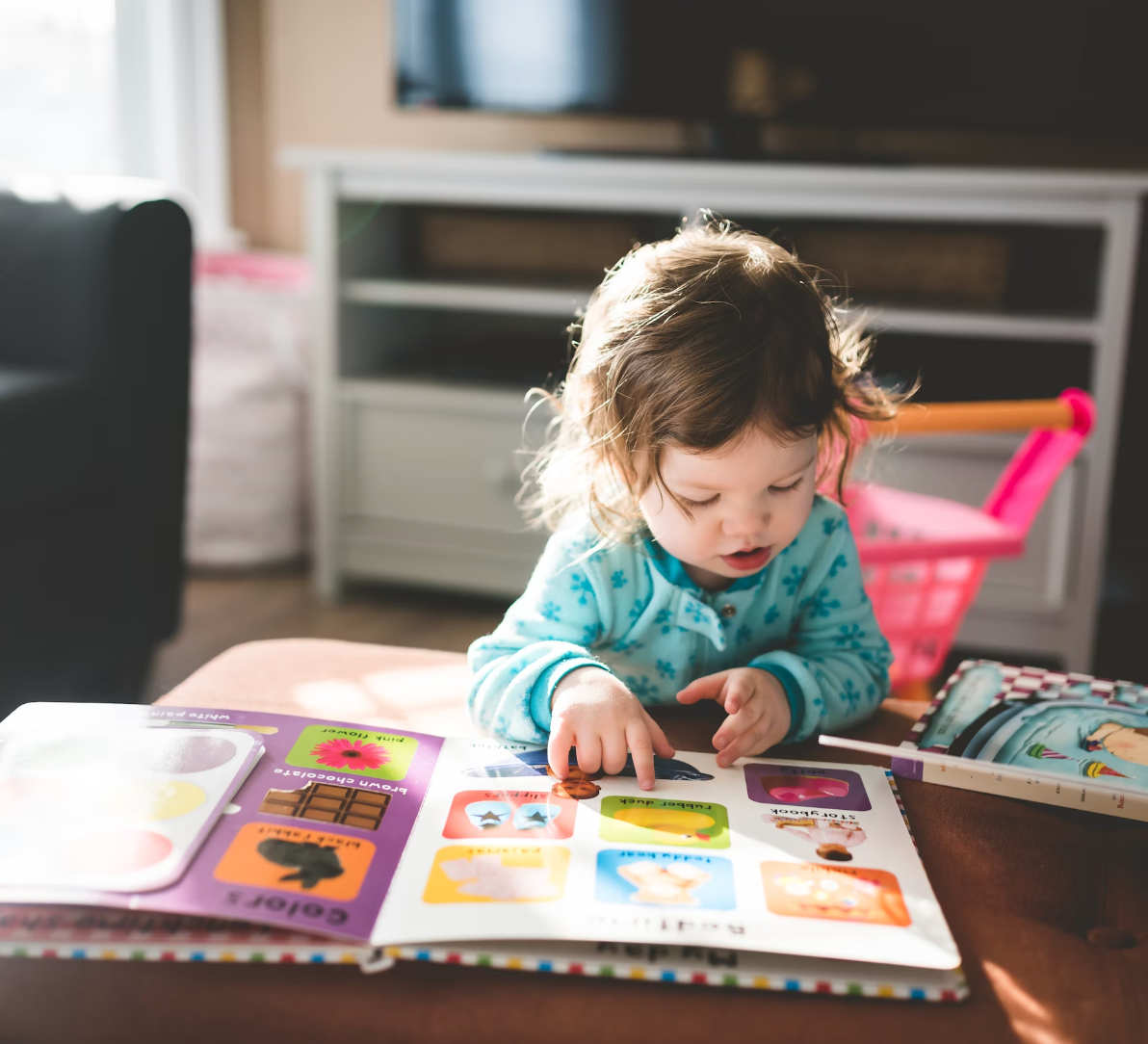 Young child pointing at pictures in a colorful board book while sitting at a table in a sunlit living room