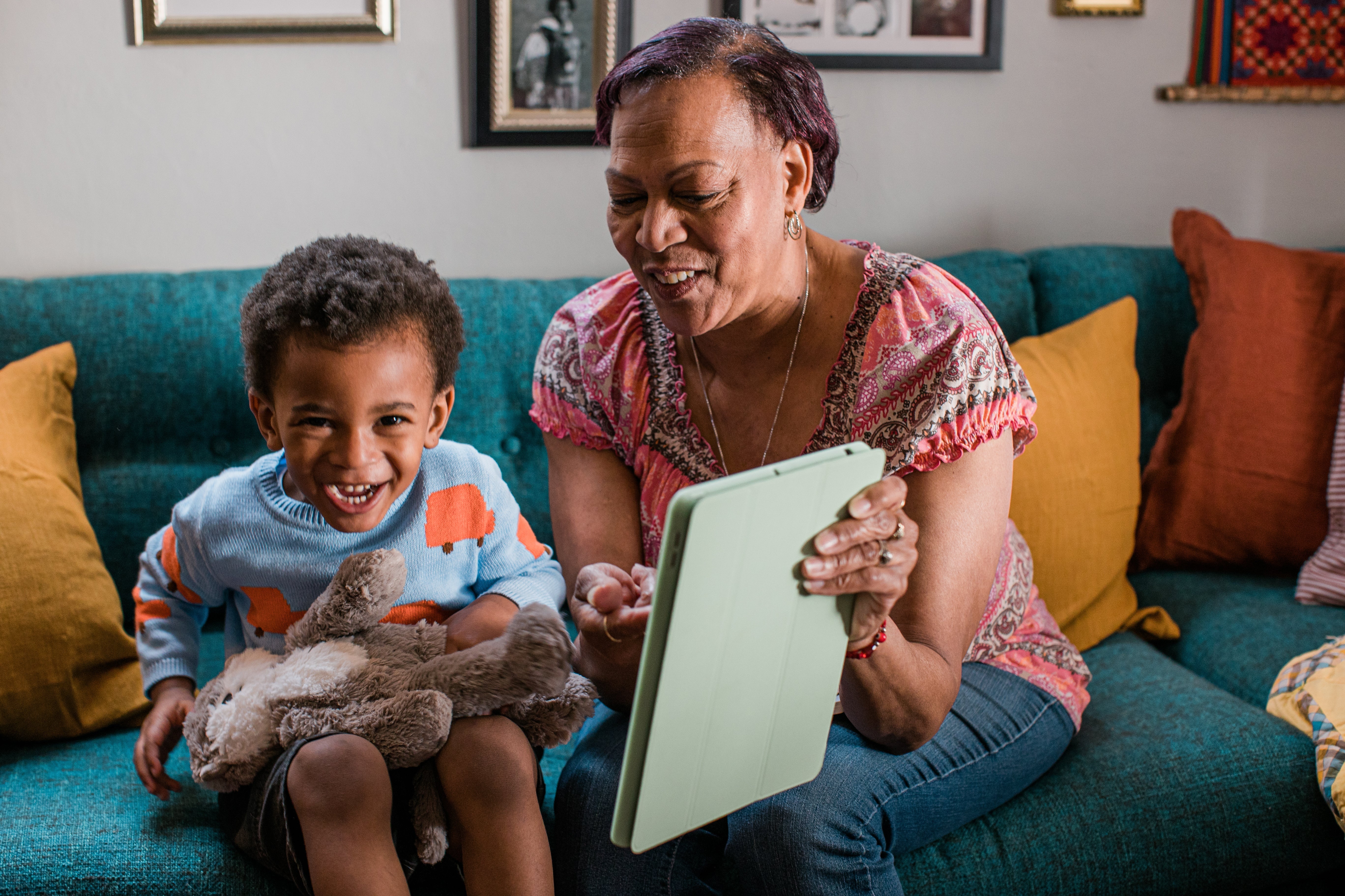 Caregiver showing a tablet to a laughing child on a teal couch while the child hugs a stuffed animal