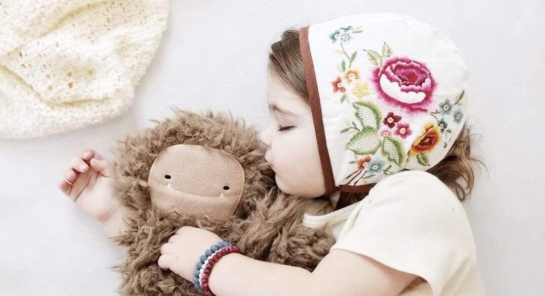 Young child wearing an embroidered floral bonnet hugging a brown plush monster on a white bed