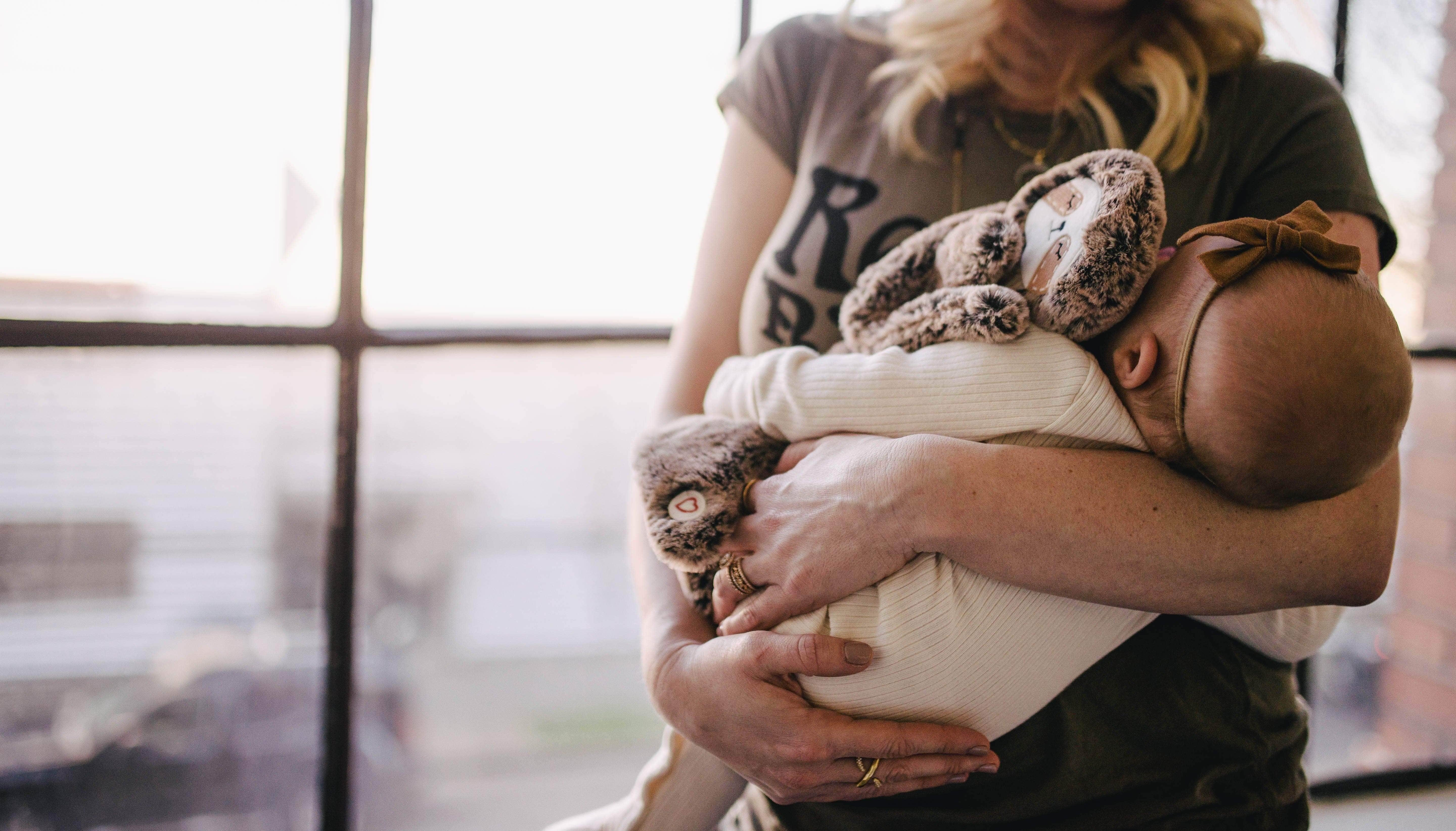 Caregiver holding a sleeping baby in a cream ribbed onesie with a brown plush bunny by a large bright window