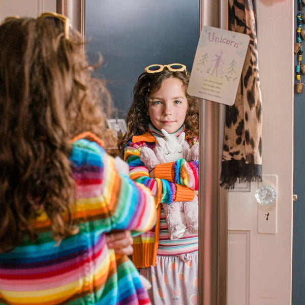 Child standing in front of mirror in brightly colored clothing, holding Unicorn Kin, looking at the Unicorn affirmation